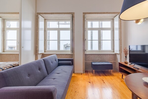 Ein modernes Wohnzimmer mit grauem Sofa, Holzboden, Fernseher und großen Fenstern.