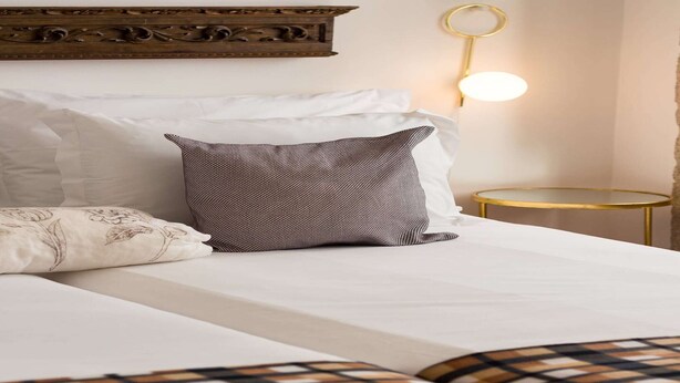 A neatly made bed with white and patterned pillows, a small round table with a gold frame, and a wall-mounted light fixture. - Stylish Porto studio with city views (Oporto)