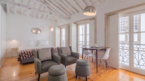 Amplia habitación con techo de madera, una cama con manta a cuadros, dos sillones grises, un pequeño reposapiés, una mesa de comedor con sillas y ventanas amplias con vista a edificios.