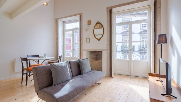 A modern living room with a grey sofa, wooden flooring, a dining area with a table and chairs, a fireplace, and large windows with a view of buildings. - Charming Porto apartment in prime location (Oporto)