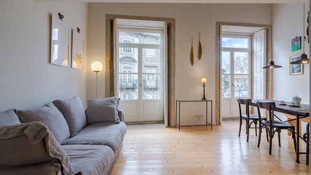 A living room with a grey sofa, wooden flooring, two windows with white frames, a small table with a lamp, and a dining area with chairs.
