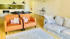A modern living room with two orange armchairs, a grey sofa, a kitchen in the background, and a small table with a lamp. - Trendy Studio in the Heart of Porto (Oporto)