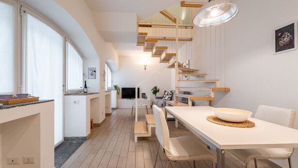 A modern kitchen with a central island, wooden staircase, and a dining area with a large table and chairs. - Viale Lecco 105 in Como (Como)