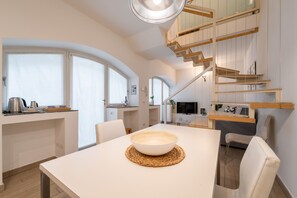 Eine moderne Küche mit Essbereich, mit einem großen weißen Tisch, einer Schüssel auf einer gewobenen Tischunterlage, weißen Stühlen und einer Holztreppe, die zu einer oberen Ebene führt.