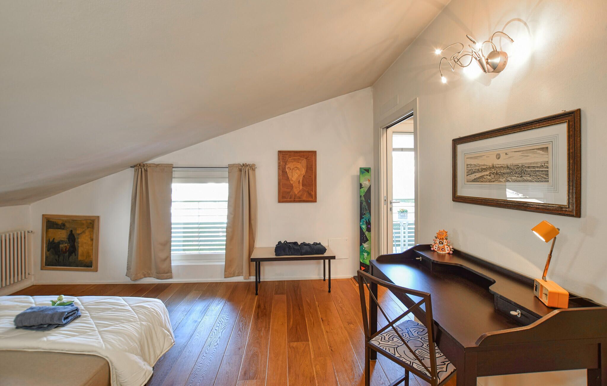 Une chambre avec un plafond en pente, un lit, un bureau et une chaise. Il y a deux fenêtres avec des rideaux, un radiateur et des œuvres d’art accrochées aux murs.