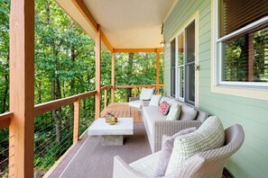 A covered porch with a sofa and chairs, a coffee table, and a view of a forested area.