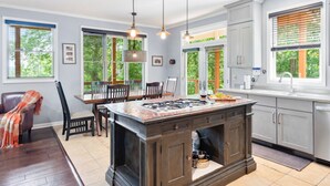 A modern kitchen with a central island, pendant lighting, and large windows with blinds. - Point of View (Fairview)