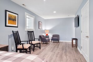 A room with a bed, two armchairs, a side table with a lamp, and a TV on the wall.