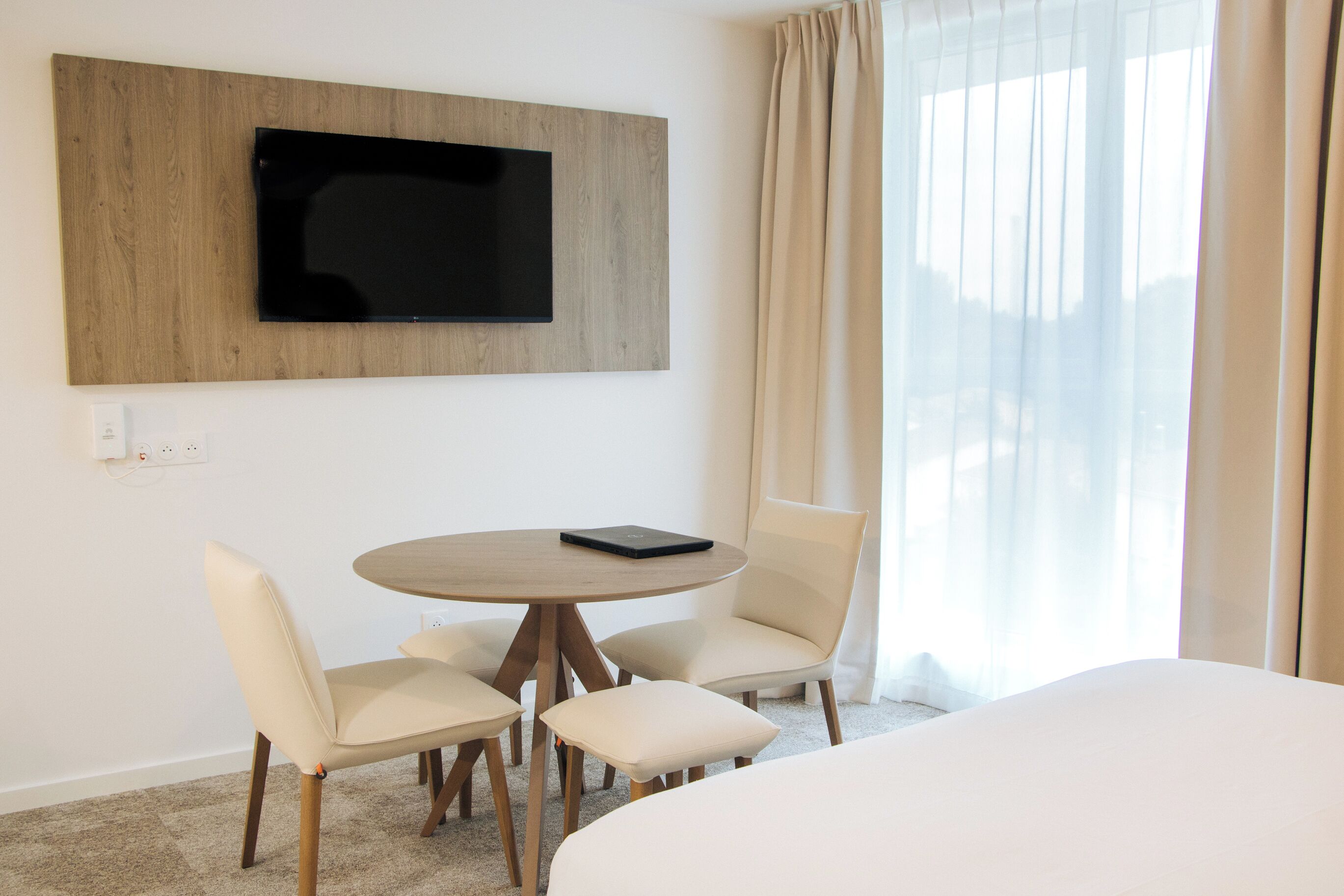 A hotel room with a bed, a round table with two chairs, a flat-screen TV mounted on a wooden panel, and a large window with sheer curtains.