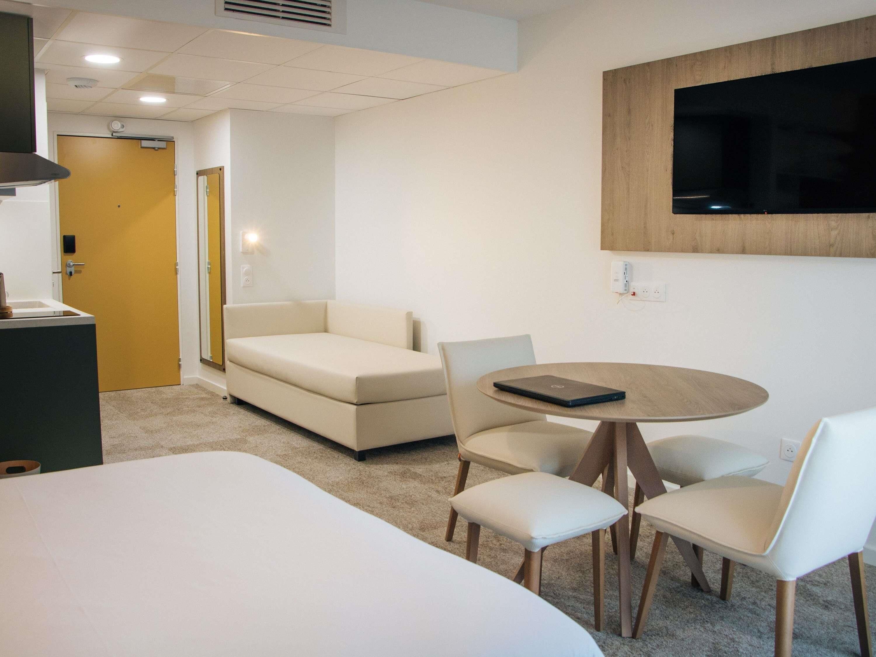 A modern hotel room with a large bed, a sofa, a dining table with chairs, a TV mounted on the wall, and a door leading to another room.