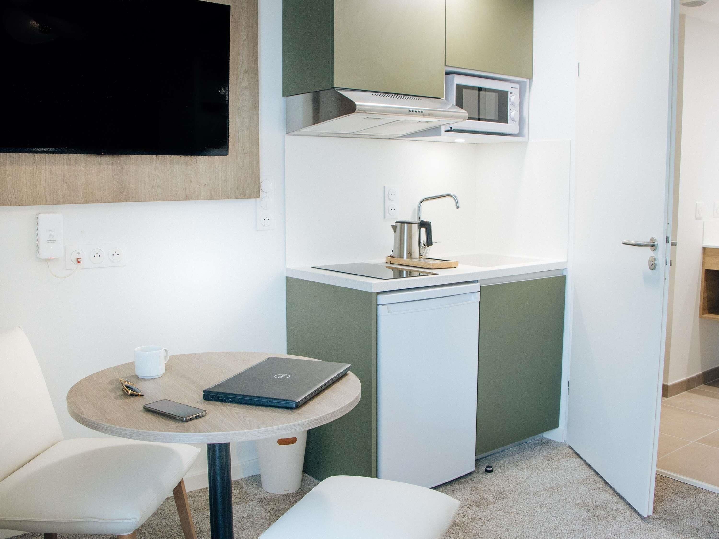 A compact kitchenette with a microwave, kettle, and sink. There is a small round table with a laptop and a cup.