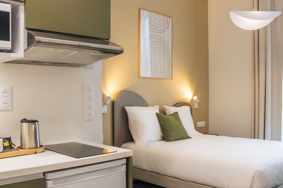A hotel room with a bed, bedside table, kettle, and a wall-mounted light fixture.
