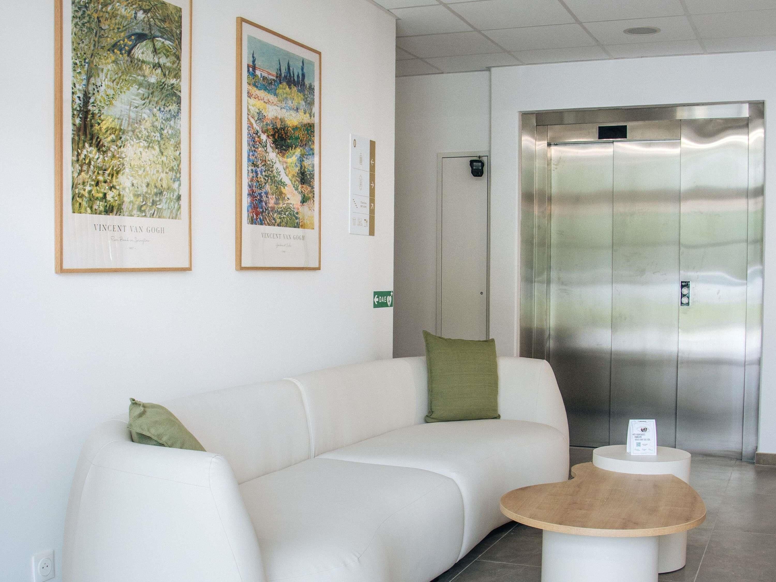Un espace d’accueil avec un canapé blanc, des coussins verts, deux tableaux encadrés, une petite table en bois et un ascenseur moderne.