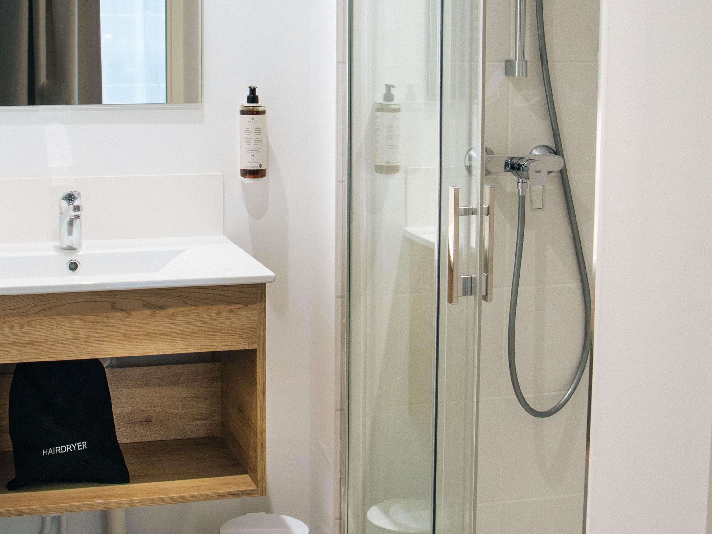 Een badkamer met een douchecabine van glas, een witte wastafel en een houten plank.