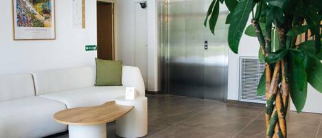 A reception area with a white sofa, a wooden coffee table, a potted plant, an elevator, and a framed artwork on the wall.