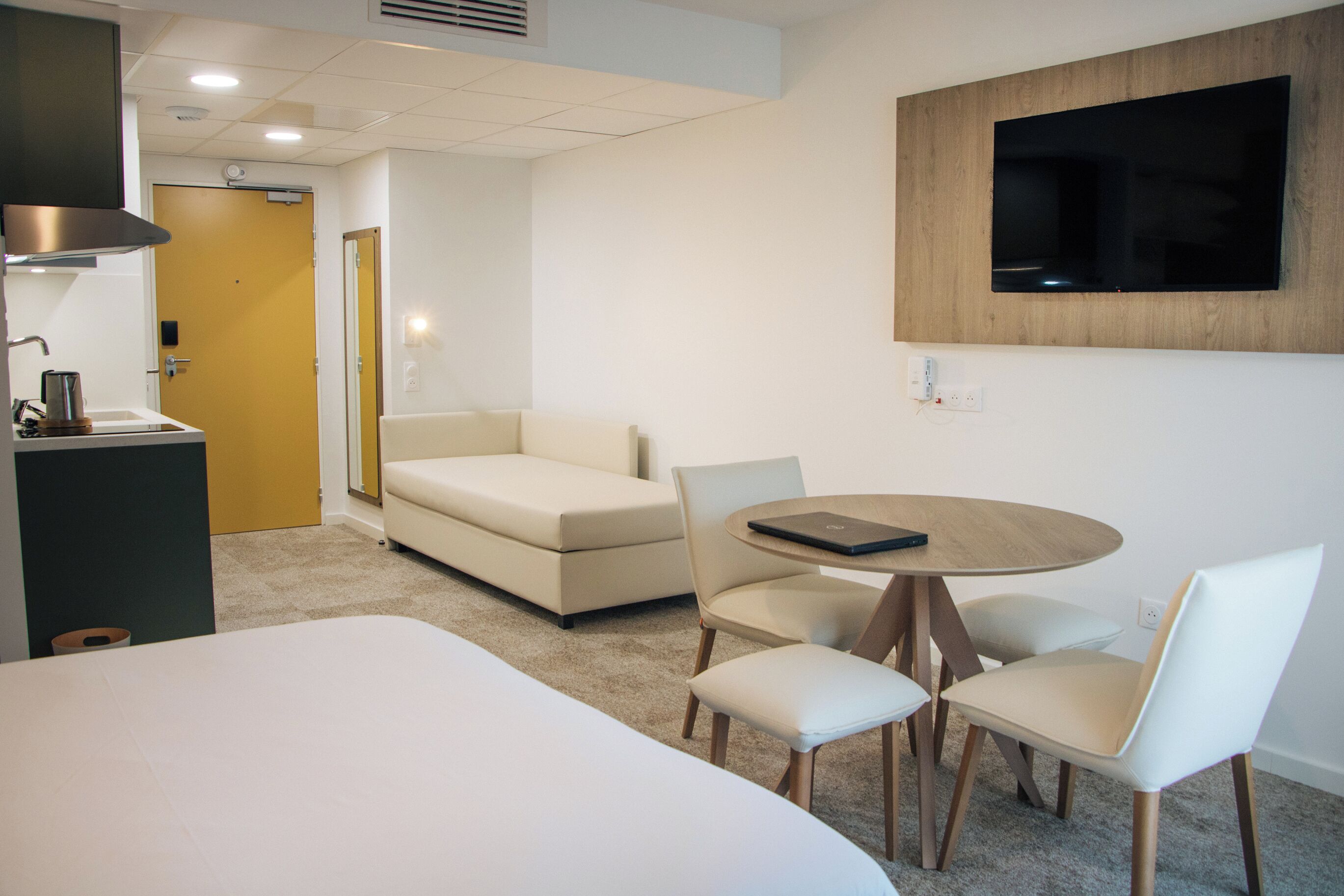 A hotel room with a bed, a sofa, a dining table with chairs, a TV mounted on the wall, and a small kitchenette area.