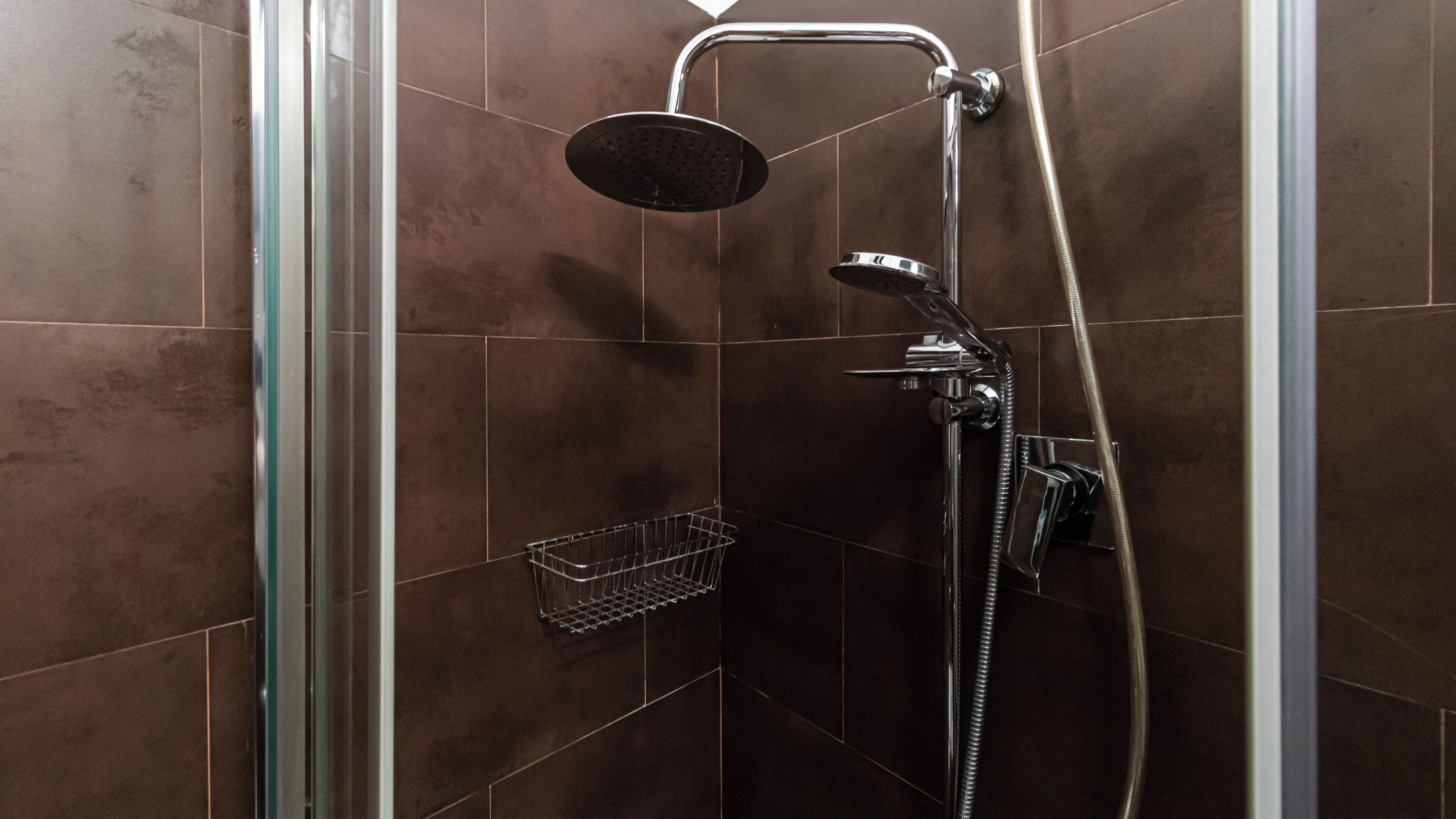 Ducha con cabezal de ducha de mano y grifo empotrado en la pared, una pequeña cesta metálica y una puerta de cristal.