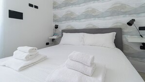 A neatly made bed with folded white towels, a grey headboard, and a wall with a wave pattern design. - Italianway Easy - Ticinese 83, 15 min to Linate (Milan)