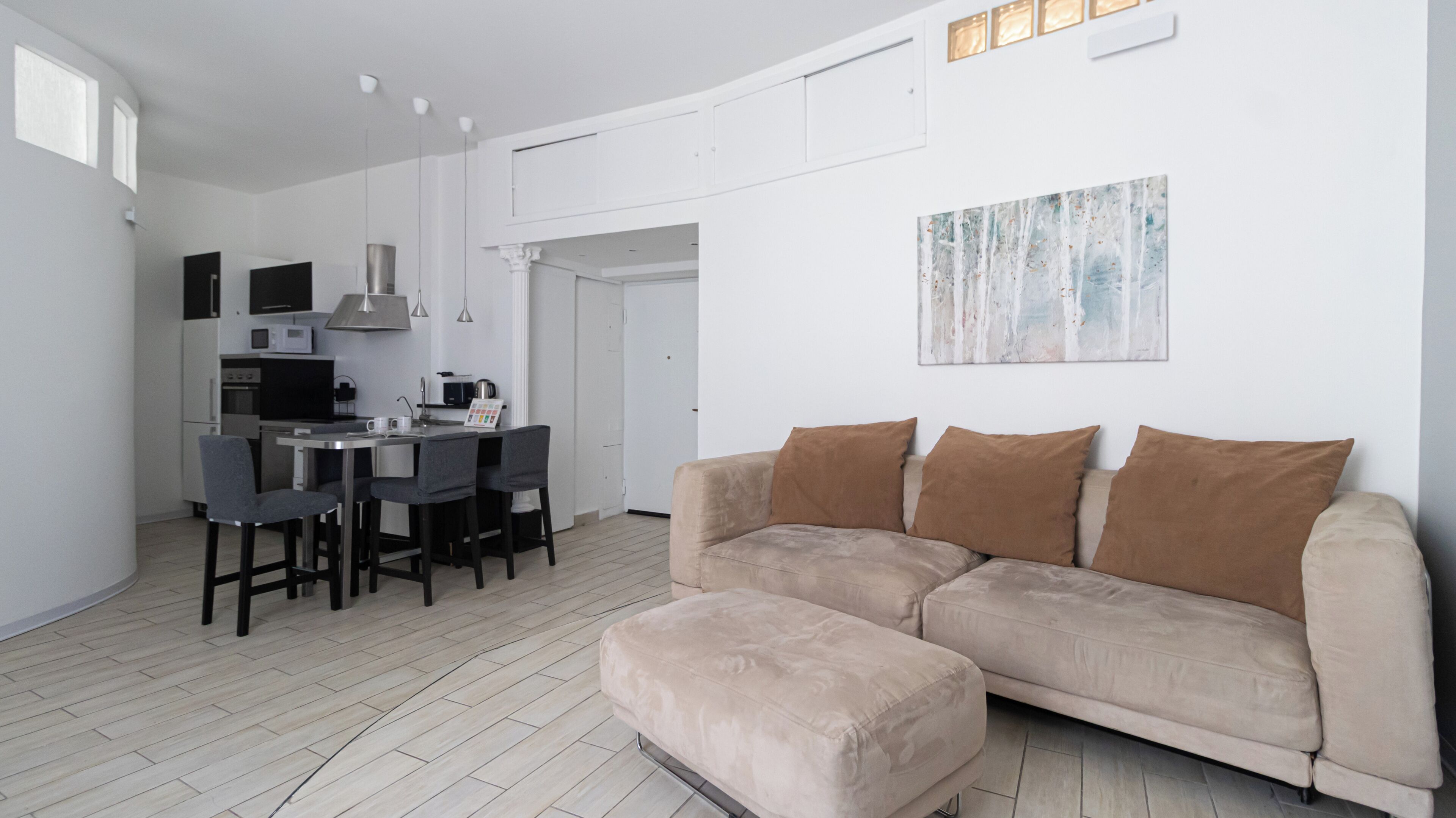 A modern living room with a beige sofa, a dining area with a small table and chairs, and a kitchen with white cabinets and appliances.