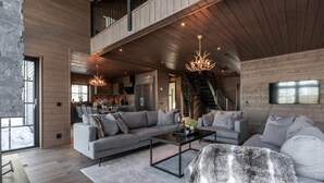 Un vaste espace de vie comprenant un grand canapé, une table basse et un lustre. Un escalier mène à l’étage supérieur, et un téléviseur est fixé au mur.