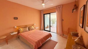 Een slaapkamer met een bed, een houten nachtkastje, een bureau met stoelen, een plafondventilator en een glazen schuifdeur die leidt naar een balkon.