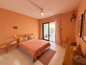 A bedroom with a bed, a wooden nightstand, a desk with chairs, a ceiling fan, and a sliding glass door leading to a balcony.