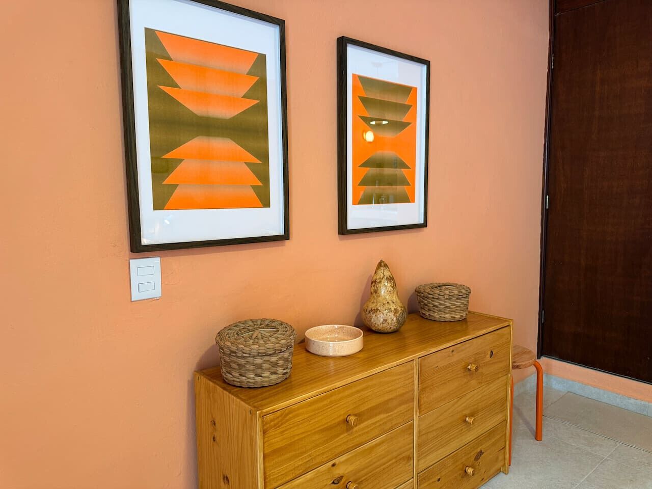 Une commode en bois, agrémentée de paniers tressés et d’un vase, est placée devant un mur de couleur pêche. Deux tableaux abstraux encadrés sont accrochés au-dessus.