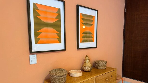 Een houten dressoir met gevlochten manden en een vaas, tegen een perzikkleurige muur, met twee ingelijste abstracte schilderijen erboven.