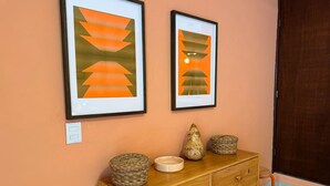 Une commode en bois, agrémentée de paniers tressés et d’un vase, est placée devant un mur de couleur pêche. Deux tableaux abstraux encadrés sont accrochés au-dessus.