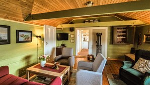 Un salon chaleureux avec un plafond en bois, des murs verts et un téléviseur. On y trouve deux canapés, une table basse et une cheminée.