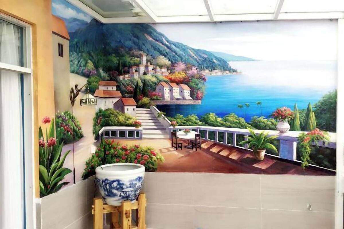 A room with a mural of a coastal village, a balcony with a table and chairs, and a large ceramic pot on a stand.