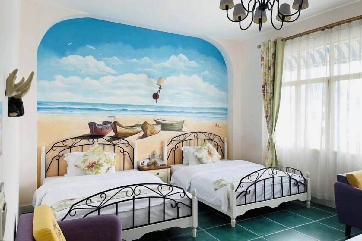 A room with two beds, a mural of a beach scene, a chandelier, and a window with curtains.
