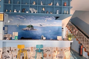 Una habitación con un mural de un faro y el mar, una escalera azul y una pared con recortes de periódico.