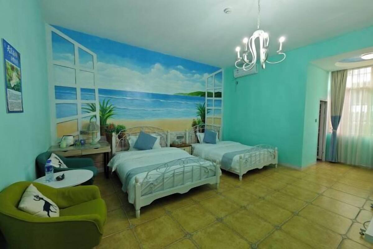 A room with two beds, a green armchair, a small table, and a beach mural on the wall.