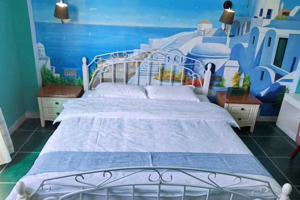 A bed with a metal frame and white bedding, a nightstand with a lamp, and a mural of a coastal scene on the wall.