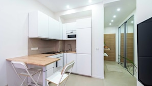 Une cuisine compacte avec des meubles blancs, un plan de travail en bois et un petit coin repas équipé d’une chaise pliante.