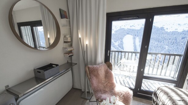 Ein Zimmer mit großem Fenster, Spiegel, Stuhl und kleinem Tisch mit Korb.