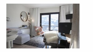 Ein modernes Hotelzimmer mit einer grauen Couch, einem runden Spiegel, einem kleinen runden Tisch mit einer Pflanze und Blick auf verschneite Berge durch das Fenster.