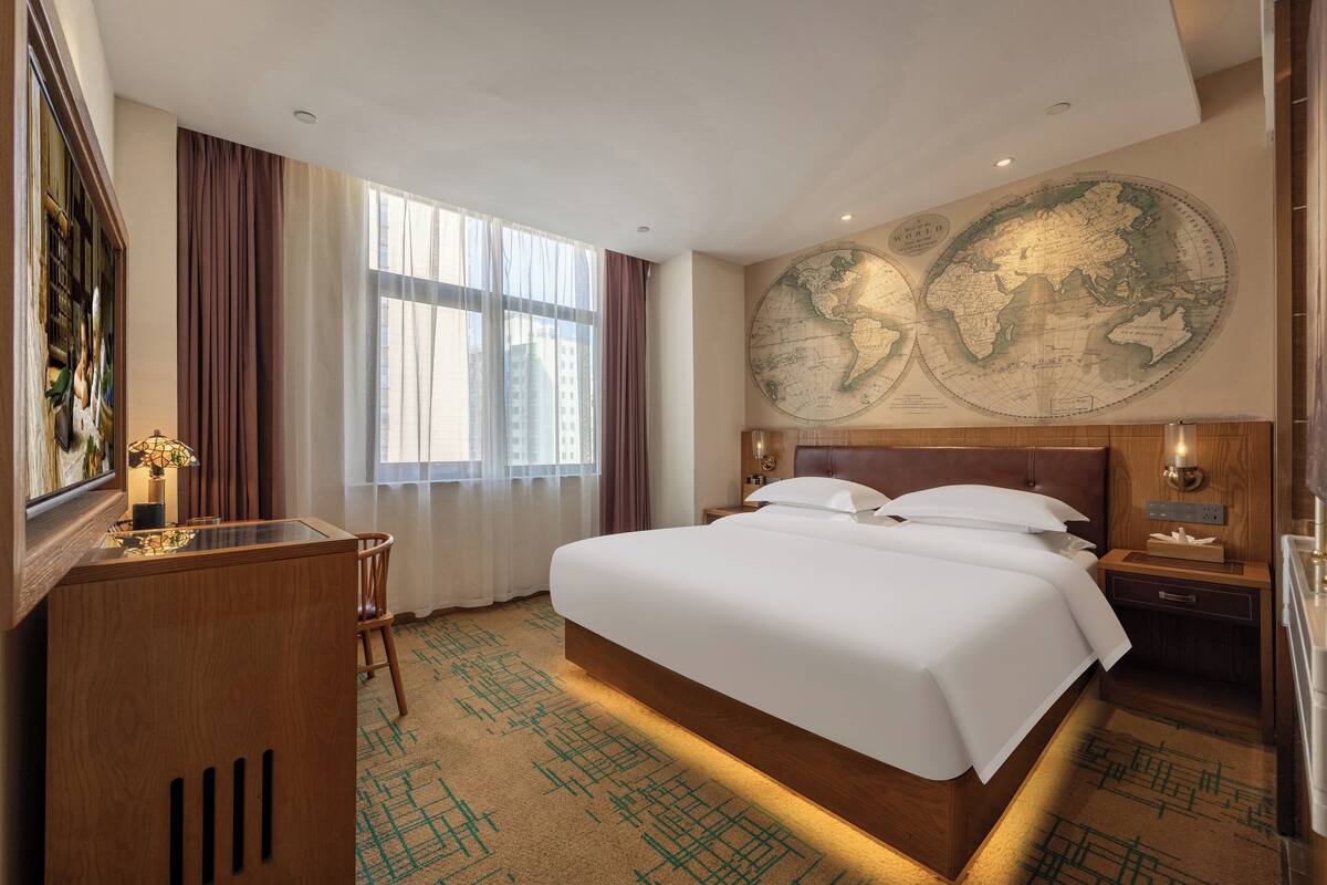 Une chambre d’hôtel avec un grand lit, un bureau avec une lampe, une chaise, une table de chevet en bois et une carte du monde accrochée au mur.