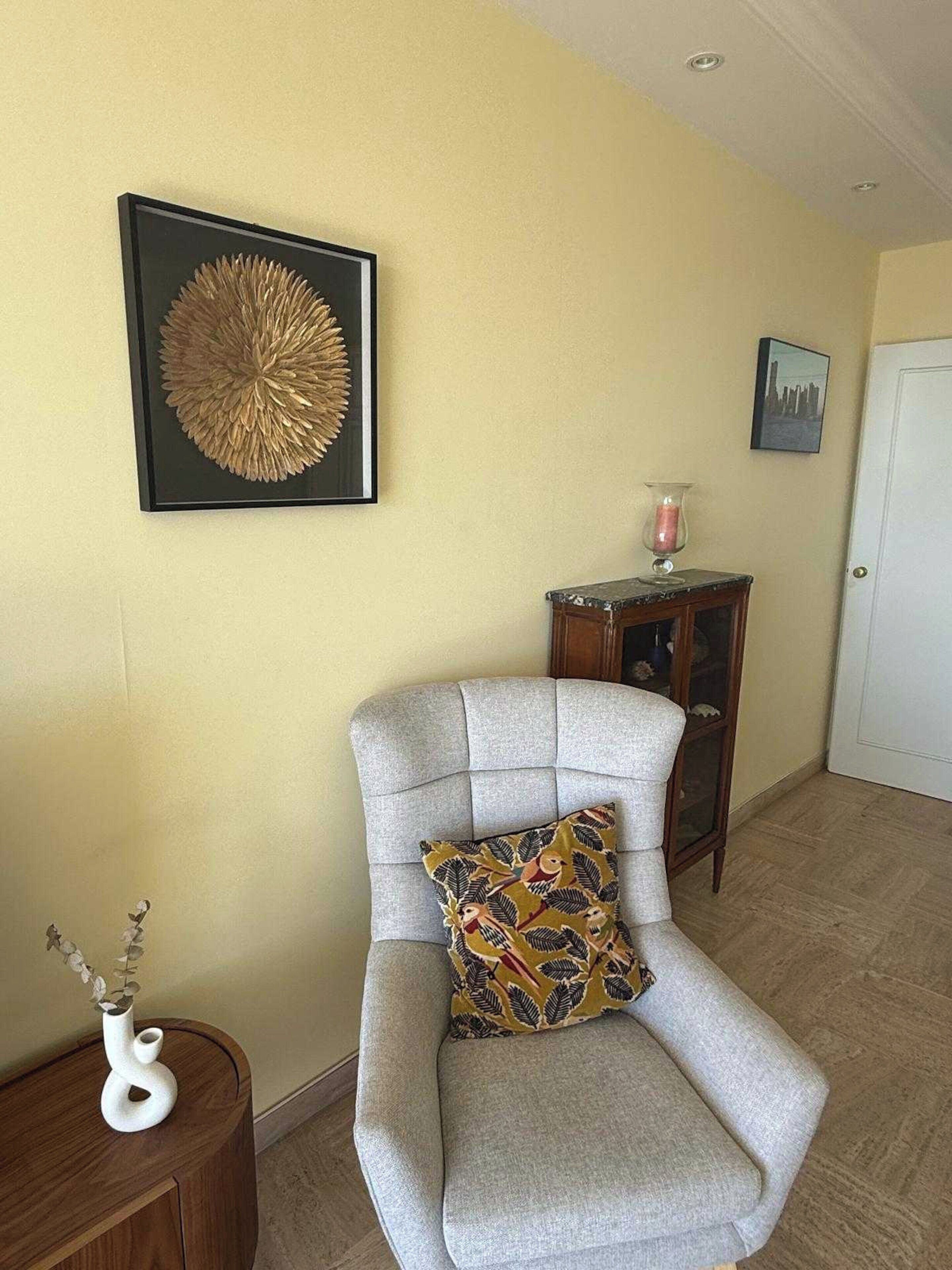 Une pièce avec un fauteuil gris, une œuvre d’art encadrée accrochée au mur, une petite table d’appoint en bois sur laquelle se trouve une bougie, et une porte visible au fond.