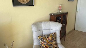 Een kamer met een grijze fauteuil, een gekaderd kunstwerk aan de muur, een klein houten bijzettafeltje met een kaars, en een deur op de achtergrond.