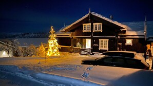 Schneebedeckte Landschaft mit einem Holzhaus, das einen Balkon und eine beleuchtete Weihnachtstanne hat.