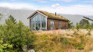A modern wooden cabin with large glass windows, surrounded by natural vegetation and a mountainous backdrop.