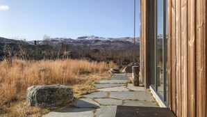 A stone pathway leading to a wooden building with large windows, surrounded by dry grass and mountains in the background.