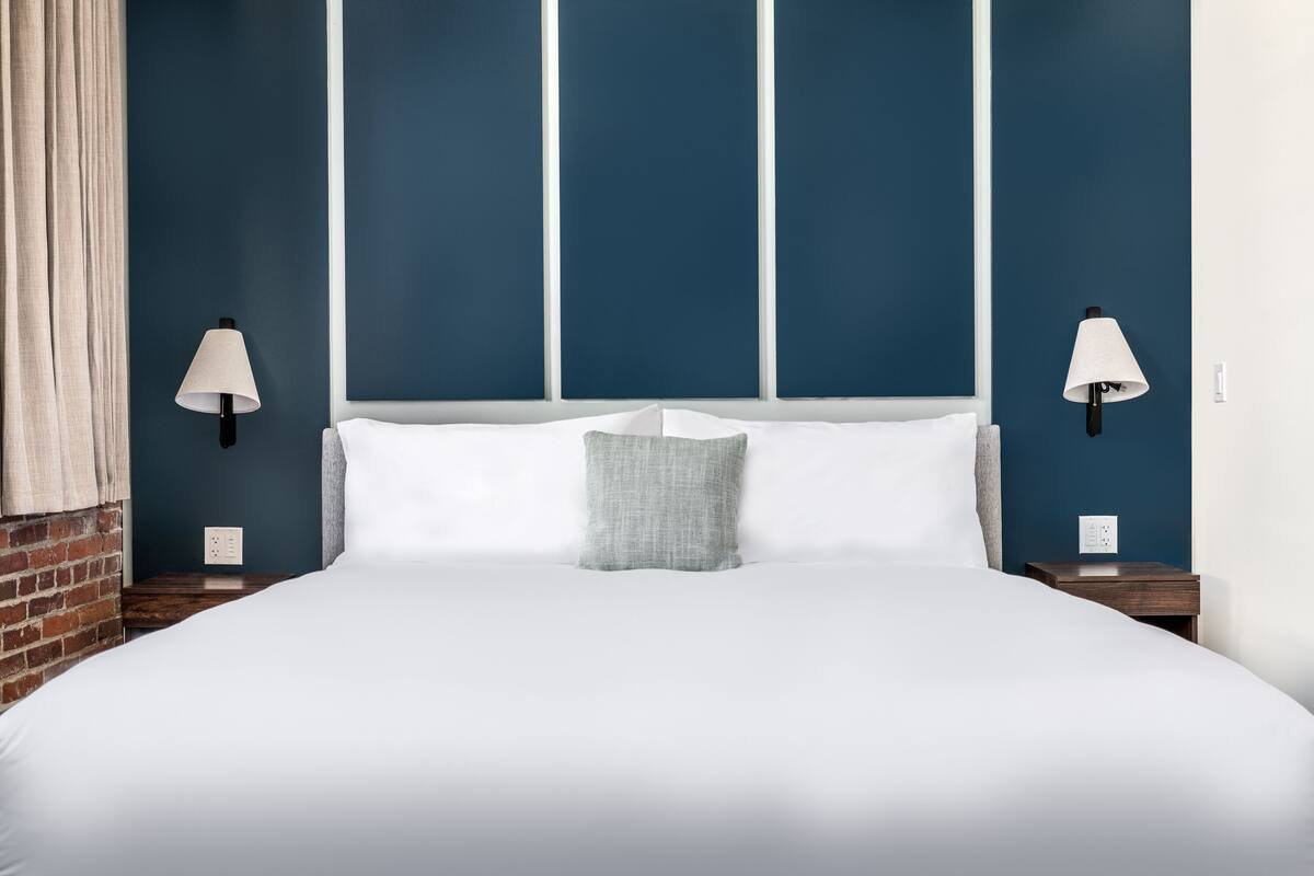 Una cama bien tendida con sábanas blancas y almohada gris, flanqueada por dos mesitas de noche con lámparas, en una habitación con paredes azules y una pared de ladrillo decorativa.