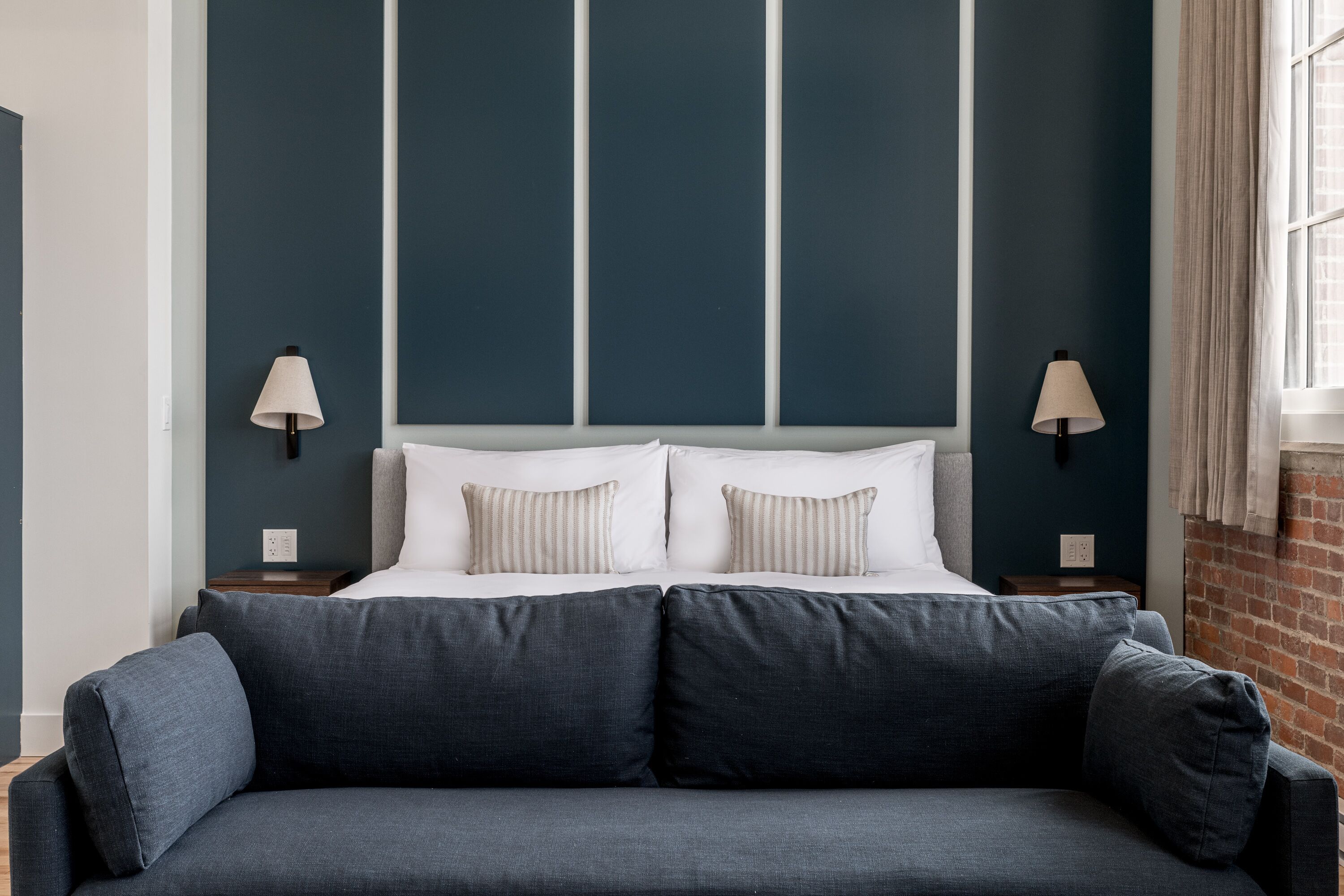 A modern hotel room with a large bed, two bedside lamps, a grey sofa, and a brick wall.