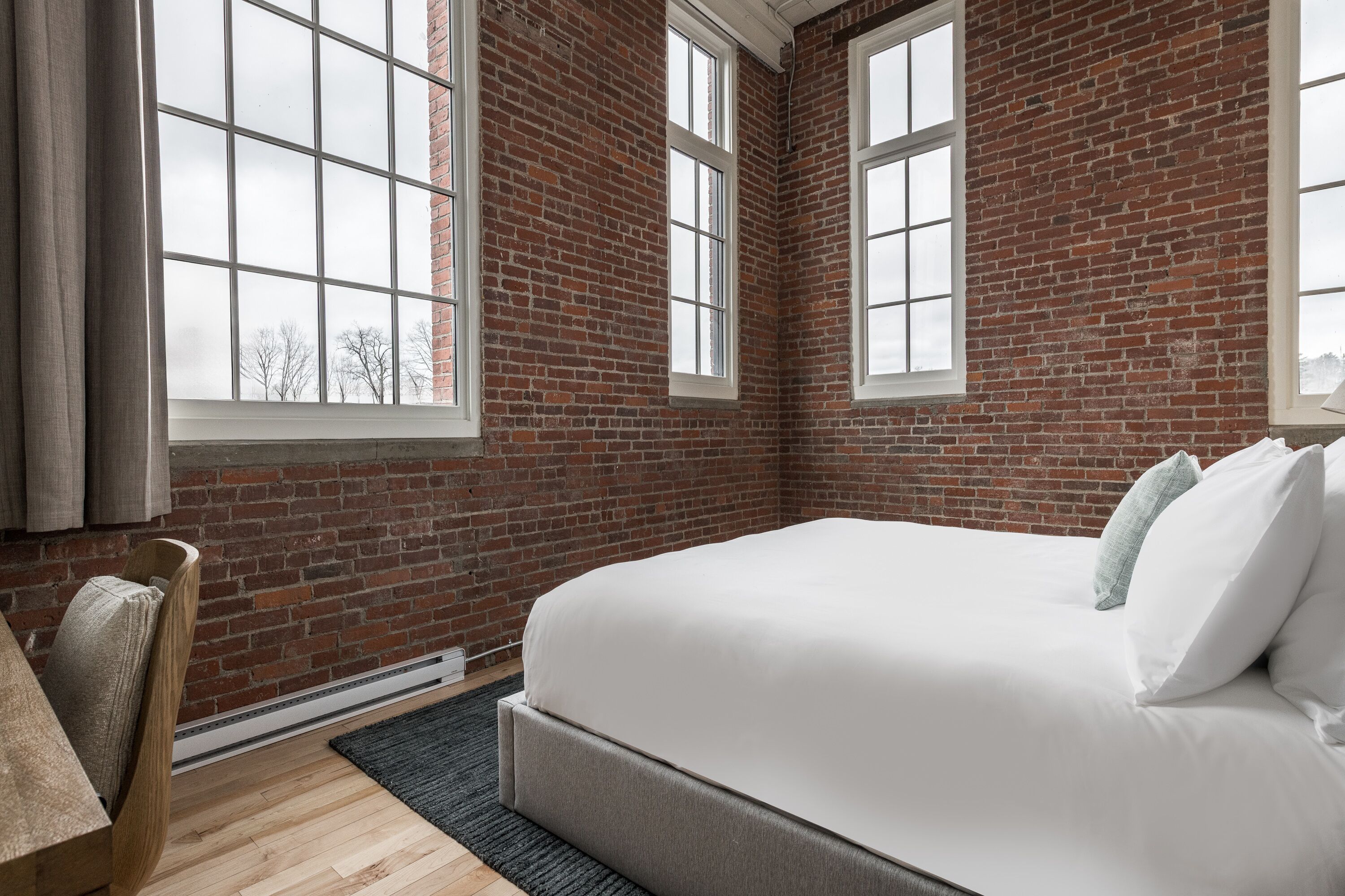 Un dormitorio con pared de ladrillo, ventanas amplias, una cama y una silla.