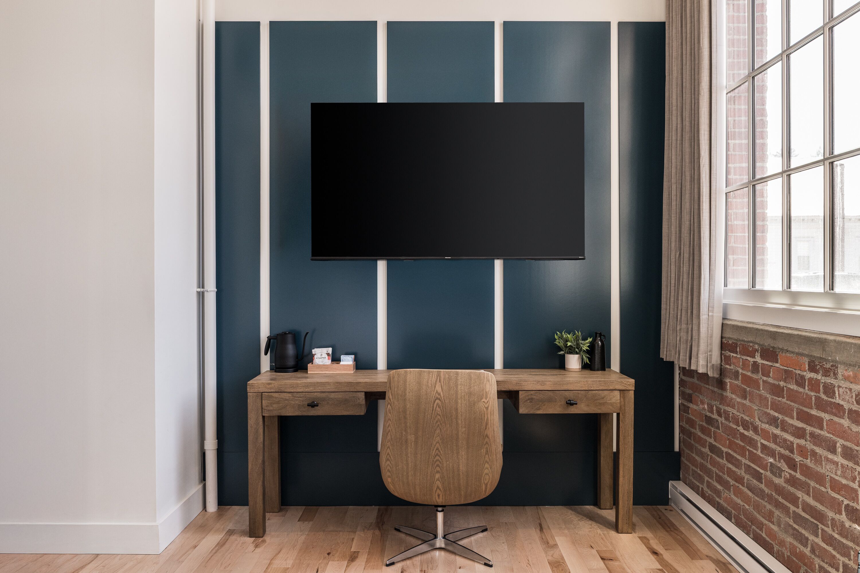 A modern office space with a large flat-screen TV mounted on the wall, a wooden desk with two drawers, and a small potted plant on the desk.