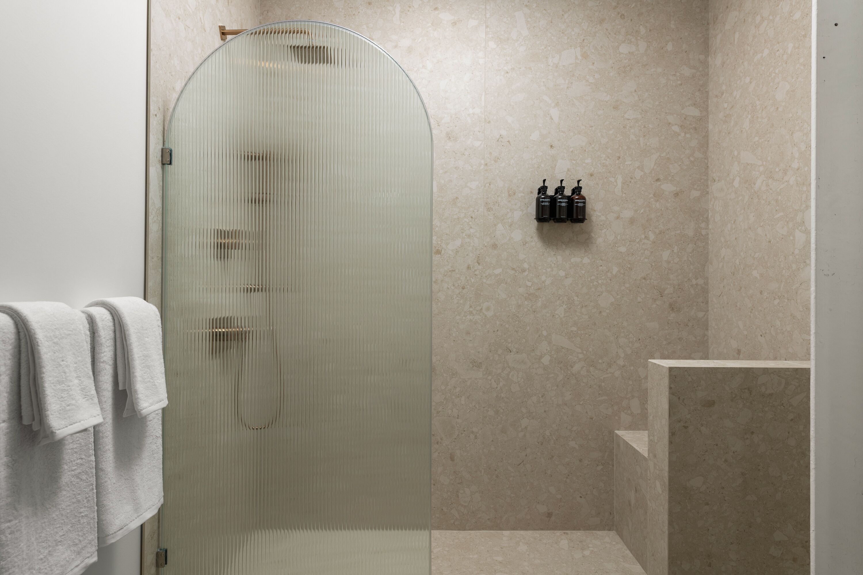 A modern bathroom with a glass shower enclosure, a marble wall, and a shelf with toiletries.