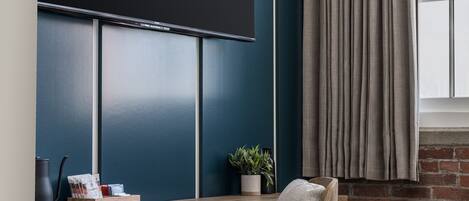 A modern office space with a dark blue panel wall, a wooden desk, a grey upholstered chair, and a large flat-screen TV mounted above the desk.